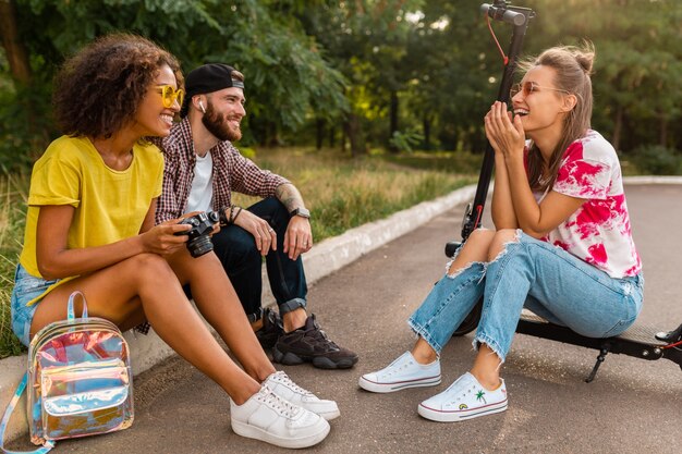 Feliz joven compañía de amigos sonrientes sentados en el parque sobre el césped con patinete eléctrico, hombre y mujer divirtiéndose juntos