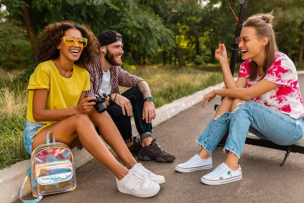 Feliz joven compañía de amigos sonrientes sentados en el parque sobre el césped con patinete eléctrico, hombre y mujer divirtiéndose juntos