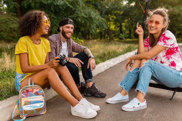 Feliz joven compañía de amigos sonrientes sentados en el parque sobre el césped con patinete eléctrico, hombre y mujer divirtiéndose juntos