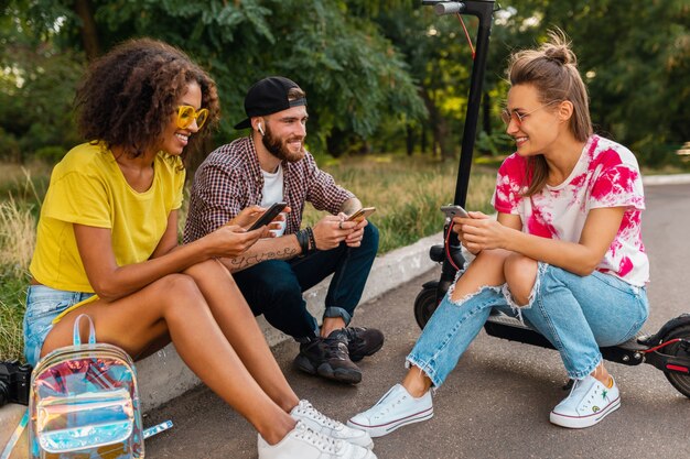 Feliz joven compañía de amigos sonrientes sentados en el parque sobre el césped con patinete eléctrico, hombre y mujer divirtiéndose juntos