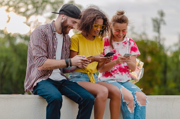 Feliz joven compañía de amigos sonrientes sentados en el parque con teléfonos inteligentes, hombres y mujeres divirtiéndose