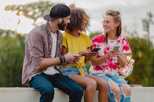 Feliz joven compañía de amigos sonrientes sentados en el parque con teléfonos inteligentes, hombres y mujeres divirtiéndose