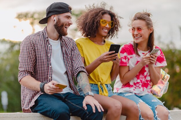 Feliz joven compañía de amigos sonrientes sentados en el parque con teléfonos inteligentes, hombres y mujeres divirtiéndose