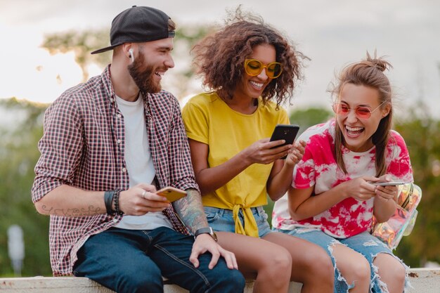 Feliz joven compañía de amigos sonrientes sentados en el parque con teléfonos inteligentes, hombres y mujeres divirtiéndose