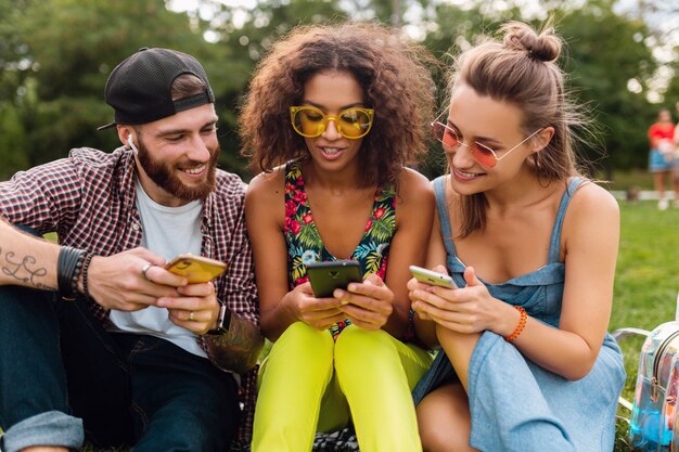 Feliz joven compañía de amigos sonrientes sentados en el parque usando teléfonos inteligentes, hombres y mujeres divirtiéndose juntos