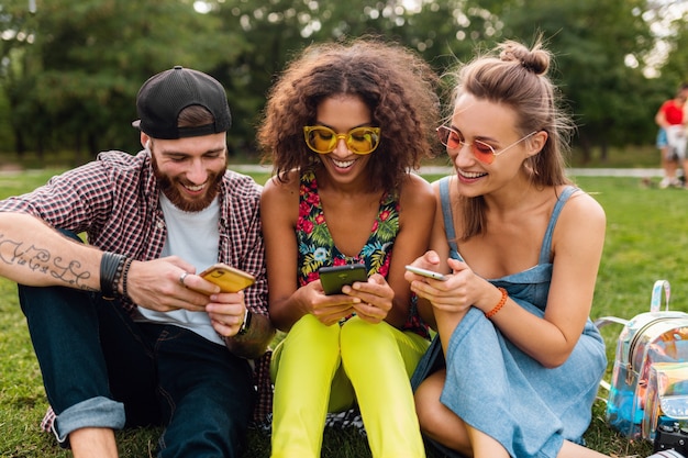 Feliz joven compañía de amigos sonrientes sentados en el parque usando teléfonos inteligentes, hombres y mujeres divirtiéndose juntos