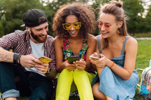 Feliz joven compañía de amigos sonrientes sentados en el parque usando teléfonos inteligentes, hombres y mujeres divirtiéndose juntos