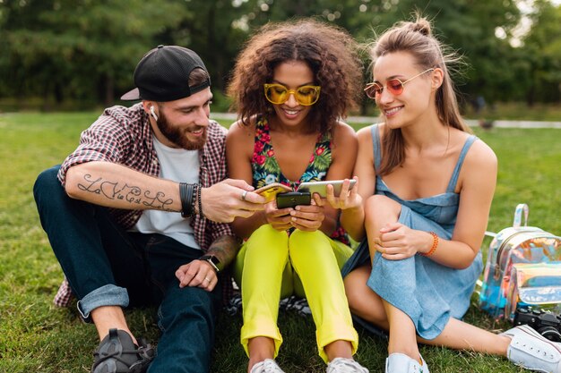 Feliz joven compañía de amigos sonrientes sentados en el parque usando teléfonos inteligentes, hombres y mujeres divirtiéndose juntos