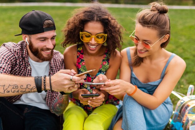 Feliz joven compañía de amigos sonrientes sentados en el parque usando teléfonos inteligentes, hombres y mujeres divirtiéndose juntos