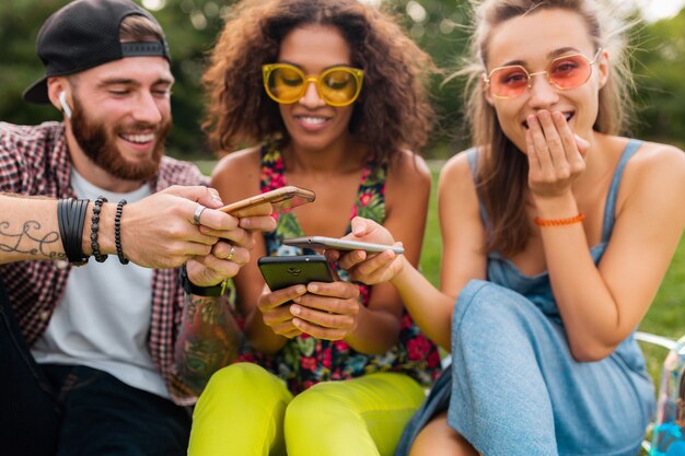 Feliz joven compañía de amigos sonrientes sentados en el parque usando teléfonos inteligentes, hombres y mujeres divirtiéndose juntos