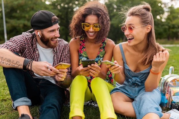 Feliz joven compañía de amigos sonrientes sentados en el parque usando teléfonos inteligentes, hombres y mujeres divirtiéndose juntos