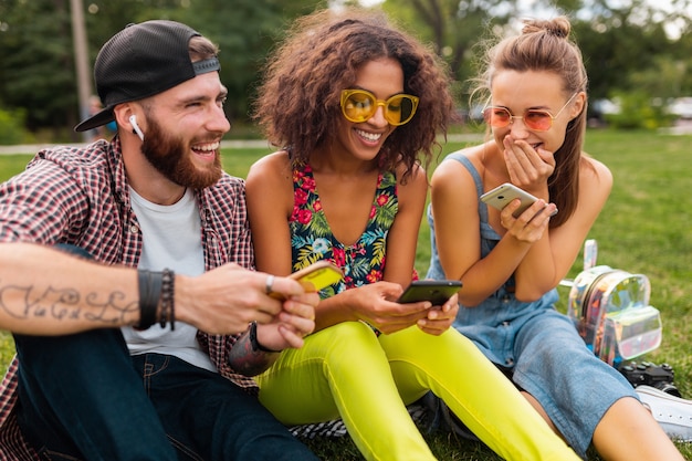 Foto gratuita feliz joven compañía de amigos sonrientes sentados en el parque usando teléfonos inteligentes, hombres y mujeres divirtiéndose juntos