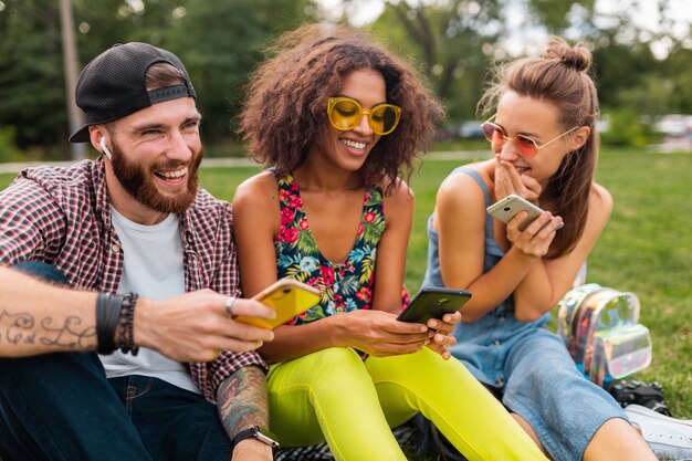 Feliz joven compañía de amigos sonrientes sentados en el parque usando teléfonos inteligentes, hombres y mujeres divirtiéndose juntos
