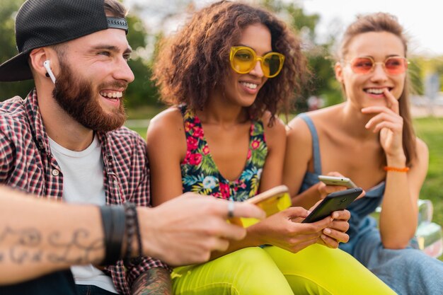 Feliz joven compañía de amigos sonrientes sentados en el parque usando teléfonos inteligentes, hombres y mujeres divirtiéndose juntos