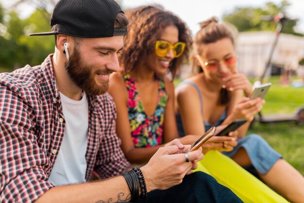 Feliz joven compañía de amigos sonrientes sentados en el parque usando teléfonos inteligentes, hombres y mujeres divirtiéndose juntos