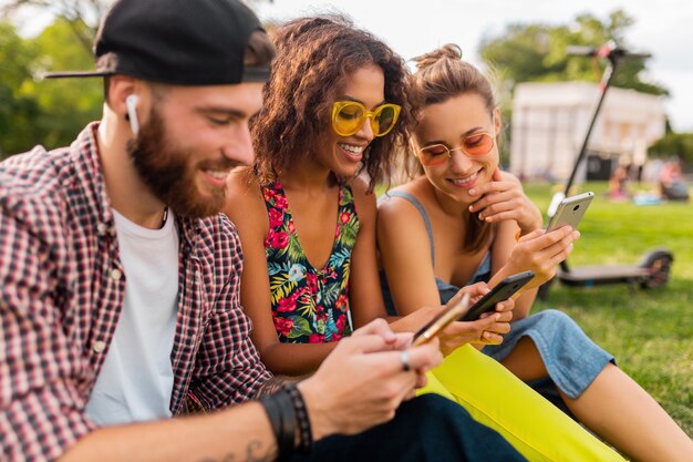 Feliz joven compañía de amigos sonrientes sentados en el parque usando teléfonos inteligentes, hombres y mujeres divirtiéndose juntos