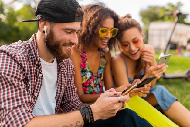 Feliz joven compañía de amigos sonrientes sentados en el parque usando teléfonos inteligentes, hombres y mujeres divirtiéndose juntos