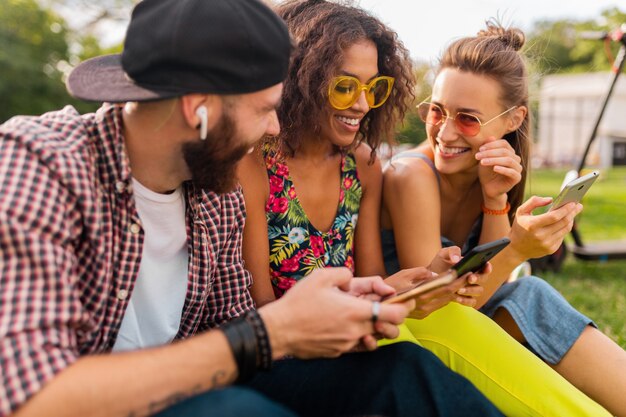 Feliz joven compañía de amigos sonrientes sentados en el parque usando teléfonos inteligentes, hombres y mujeres divirtiéndose juntos