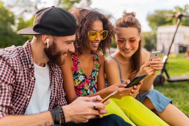 Feliz joven compañía de amigos sonrientes sentados en el parque usando teléfonos inteligentes, hombres y mujeres divirtiéndose juntos