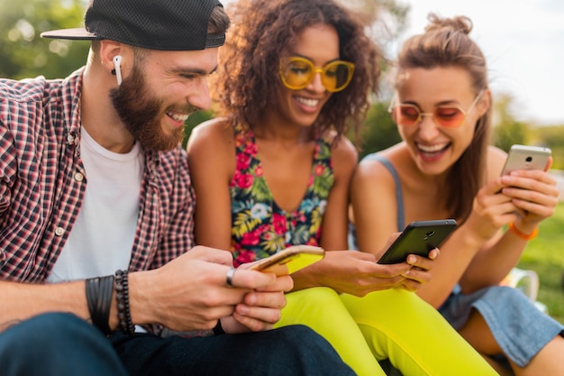 Feliz joven compañía de amigos sonrientes sentados en el parque usando teléfonos inteligentes, hombres y mujeres divirtiéndose juntos