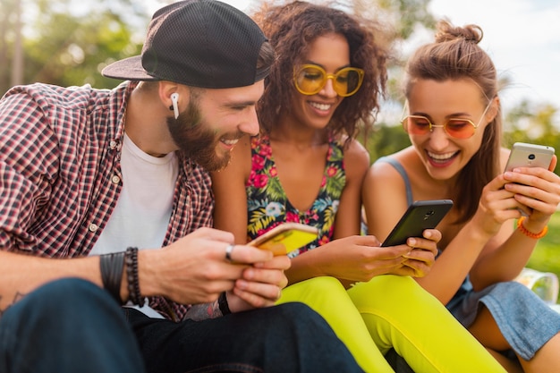 Feliz joven compañía de amigos sonrientes sentados en el parque usando teléfonos inteligentes, hombres y mujeres divirtiéndose juntos