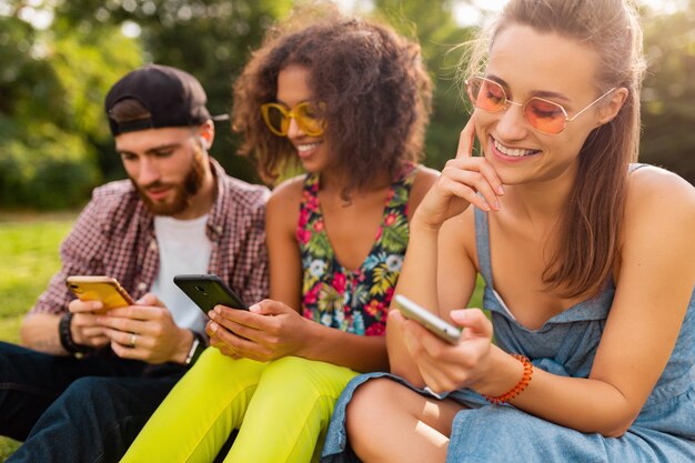 Feliz joven compañía de amigos sonrientes sentados en el parque usando teléfonos inteligentes, hombres y mujeres divirtiéndose juntos