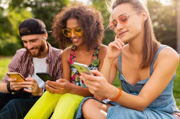 Feliz joven compañía de amigos sonrientes sentados en el parque usando teléfonos inteligentes, hombres y mujeres divirtiéndose juntos