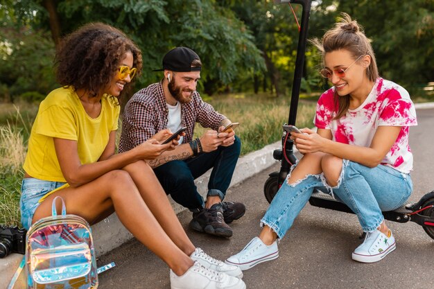 Feliz joven compañía de amigos sonrientes sentados en el parque usando teléfonos inteligentes, hombres y mujeres divirtiéndose juntos