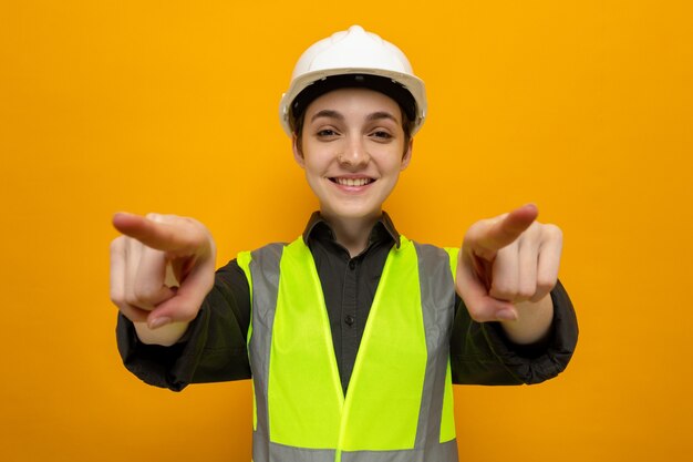 Feliz joven constructor mujer en chaleco de construcción y casco de seguridad sonriendo confiado apuntando con los dedos índices de pie en naranja