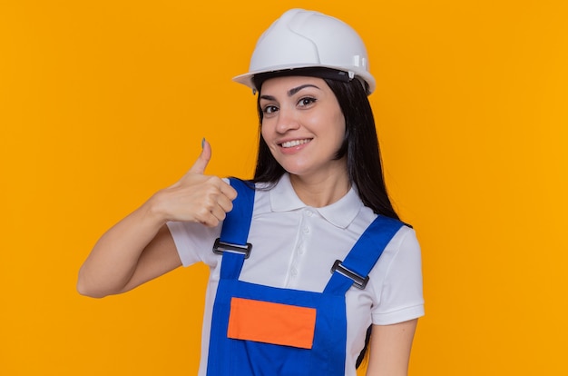 Feliz joven constructor mujer en uniforme de construcción y casco de seguridad mirando al frente sonriendo alegremente mostrando los pulgares para arriba de pie sobre la pared naranja