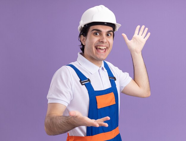Feliz joven constructor en uniforme de construcción y casco de seguridad mirando al frente gesticulando con las manos sonriendo alegremente de pie sobre la pared púrpura