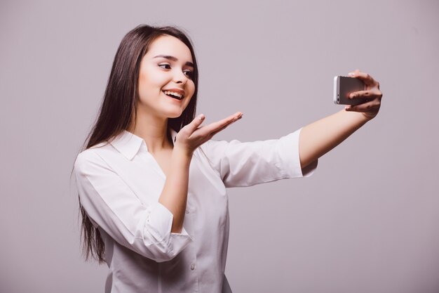 Feliz joven coqueteando tomando fotografías de sí misma en el teléfono inteligente que sopla un beso, sobre fondo blanco.