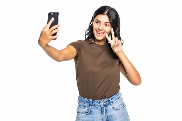 Feliz joven coqueteando tomando fotografías de sí misma en el teléfono inteligente, sobre la pared blanca