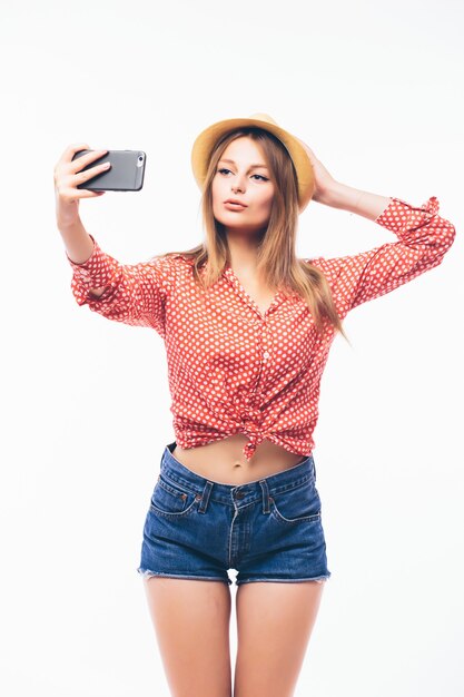 Feliz joven coqueteando tomando fotografías de sí misma a través del teléfono celular, sobre fondo blanco.