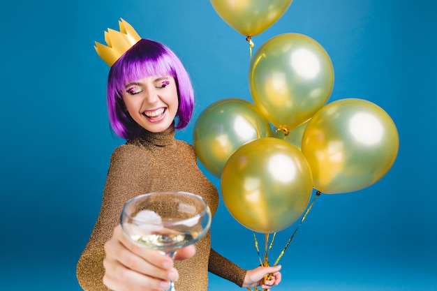 Feliz joven con corte de pelo morado, corona en la cabeza celebrando con globos dorados y champán. Vestido de lujo, fiesta de año nuevo, cumpleaños, sonriendo con los ojos cerrados.