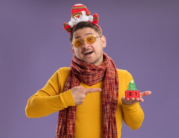 Feliz joven de cuello alto amarillo con bufanda cálida y gafas con borde divertido con santa en la cabeza mostrando cubos de juguete con fecha de año nuevo de pie sobre la pared púrpura