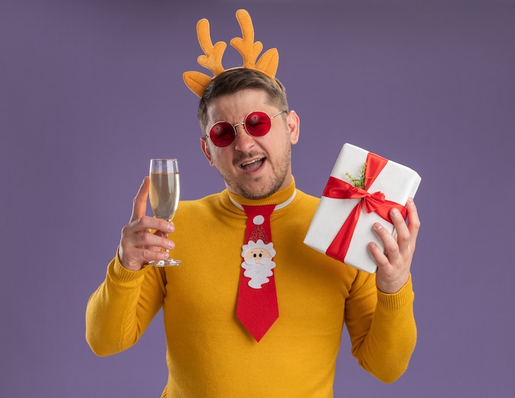Foto gratuito feliz joven con cuello alto amarillo y gafas rojas con corbata roja divertida y borde con cuernos de venado sosteniendo una copa de champán y presente smling sobre fondo púrpura