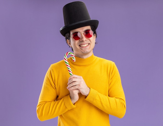 Feliz joven de cuello alto amarillo y gafas con sombrero negro sosteniendo bastón de caramelo con una sonrisa en la cara de pie sobre la pared púrpura