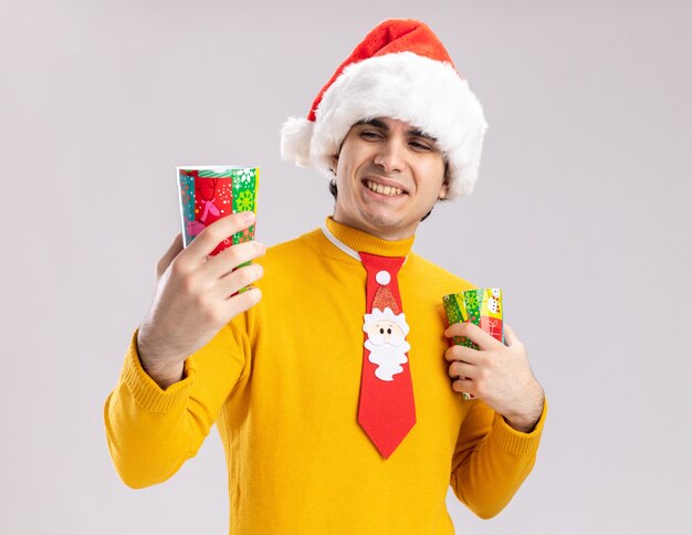 Feliz joven de cuello alto amarillo y gorro de Papá Noel con corbata divertida sosteniendo coloridos vasos de papel mirándolos sonriendo de pie sobre fondo blanco.