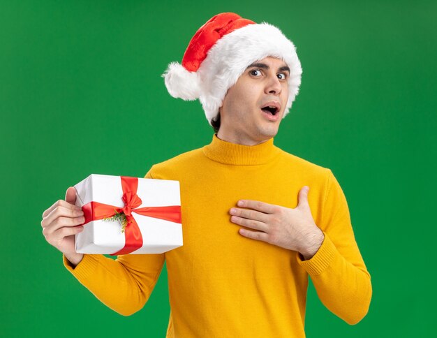 Feliz joven de cuello alto amarillo y gorro de Papá Noel con corbata divertida sosteniendo un presente mirando a la cámara sorprendido de pie sobre fondo verde