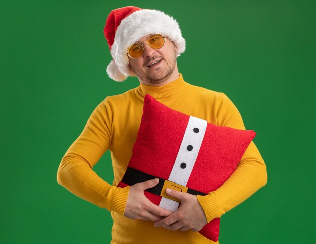 Feliz joven de cuello alto amarillo y gorro de Papá Noel con gafas sosteniendo una almohada roja divertida con una sonrisa en la cara de pie sobre la pared verde