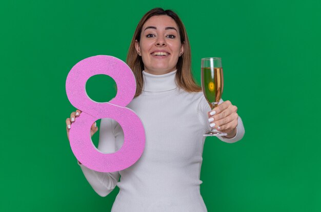 Feliz joven de cuello alto blanco sosteniendo el número ocho y una copa de champán