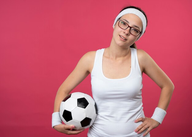 Feliz joven deportista con gafas ópticas con diadema y muñequeras pone la mano en la cintura y sostiene la bola aislada en la pared rosa