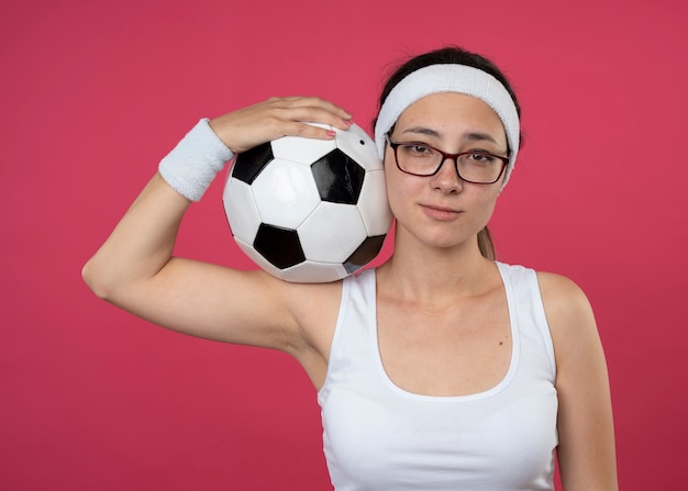 Feliz joven deportista con gafas ópticas con diadema y muñequeras sostiene la bola en el hombro