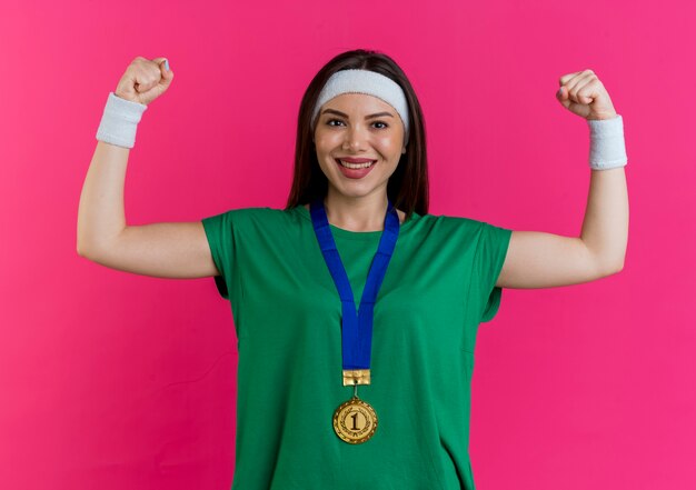 Feliz joven deportiva vistiendo diadema y muñequeras con medalla alrededor del cuello mirando haciendo gesto fuerte