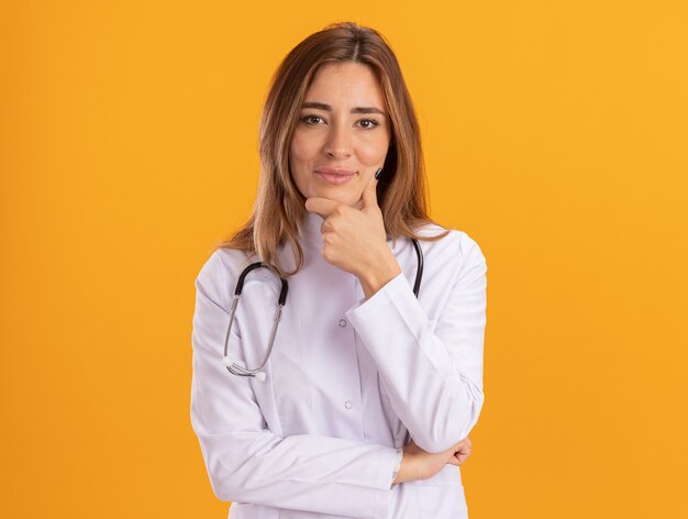 Feliz joven doctora vistiendo bata médica con estetoscopio agarró la barbilla aislada en la pared amarilla