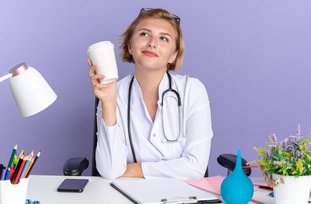 Feliz joven doctora vistiendo bata médica con estetoscopio y gafas se sienta a la mesa con herramientas médicas sosteniendo una taza de café aislada en la pared azul