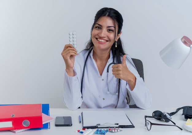 Feliz joven doctora vistiendo bata médica y estetoscopio sentado en el escritorio con herramientas médicas sosteniendo un paquete de tabletas médicas mirando mostrando pulgar hacia arriba aislado