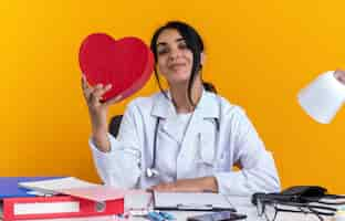 Foto gratuita feliz joven doctora vistiendo bata médica con estetoscopio se sienta a la mesa con herramientas médicas sosteniendo una caja en forma de corazón aislada en la pared amarilla