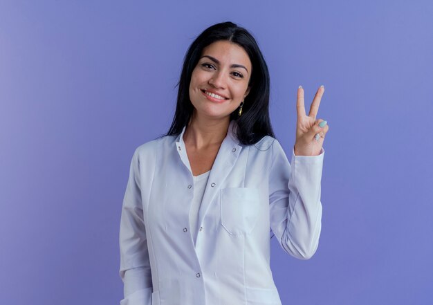 Feliz joven doctora vistiendo bata médica mirando haciendo el signo de la paz